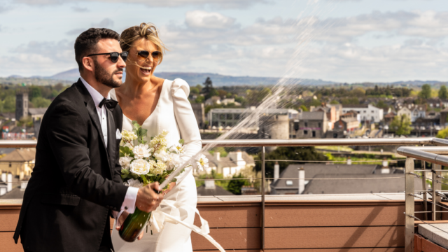 A bride and groom in formal attire stand on a rooftop, smiling as the groom pops a bottle of champagne, with city buildings and a cloudy sky in the background.