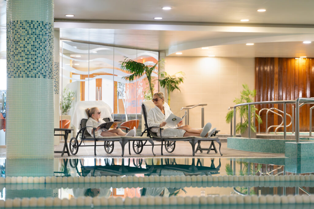 Two people in white bathrobes and slippers relax on lounge chairs by an indoor pool, reading magazines. Modern decor and potted plants set the scene for a peaceful Summer Family Break indoors.