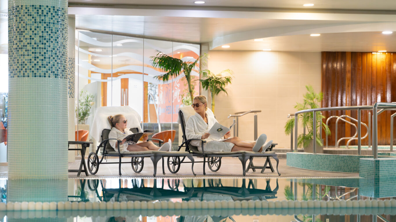 Two people in white bathrobes and slippers relax on lounge chairs by an indoor pool, reading magazines. Modern decor and potted plants set the scene for a peaceful Summer Family Break indoors.