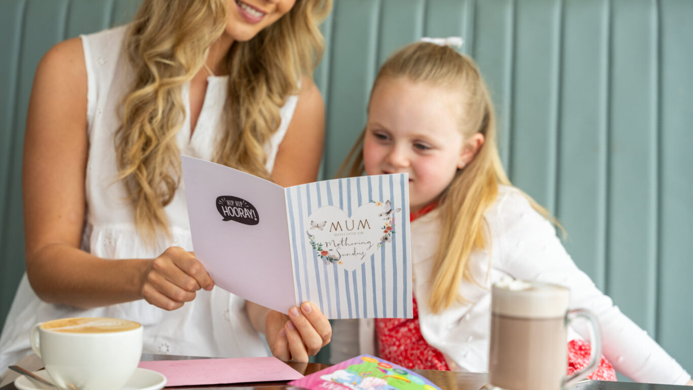 An adult and a child sit at a table, reading a card together. Drinks and colored pencils are on the table in front of them.