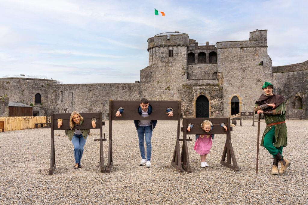 Three people pose in wooden stocks before a stone castle during their Spring Hotel Breaks, while a person dressed as a medieval character stands nearby; an Irish flag waves atop the castle.