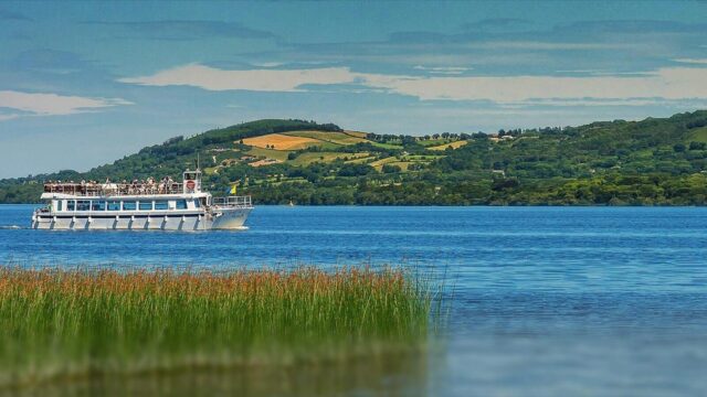 A white passenger boat travels across a blue lake with green hills and fields in the background under a partly cloudy sky.