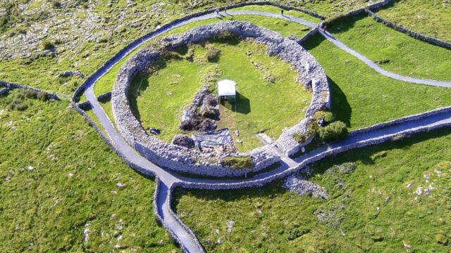 Aerial view of a large circular stone fort with grassy interior, surrounded by stone walls and pathways in a green landscape.