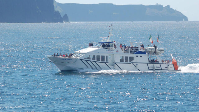 A white passenger ferry with people on board travels across a calm, blue sea with cliffs and distant land visible in the background.