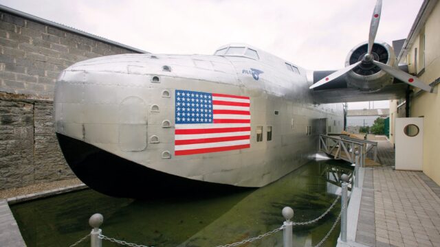 A large silver aircraft with an American flag painted on its side is displayed on a shallow pool of water between buildings.