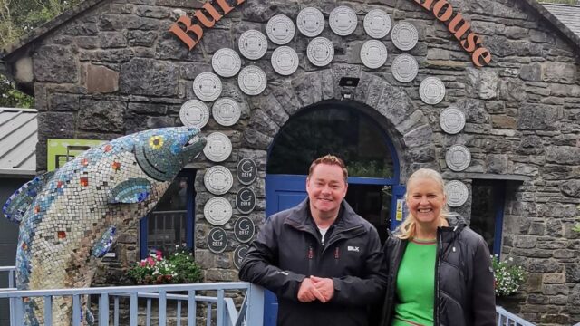 Two people stand in front of the stone building entrance to Burren Smokehouse, next to a large mosaic fish sculpture.