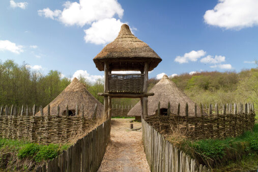 A reconstructed ancient settlement with thatched roundhouses and a wooden watchtower, surrounded by a woven fence, under a partly cloudy sky.