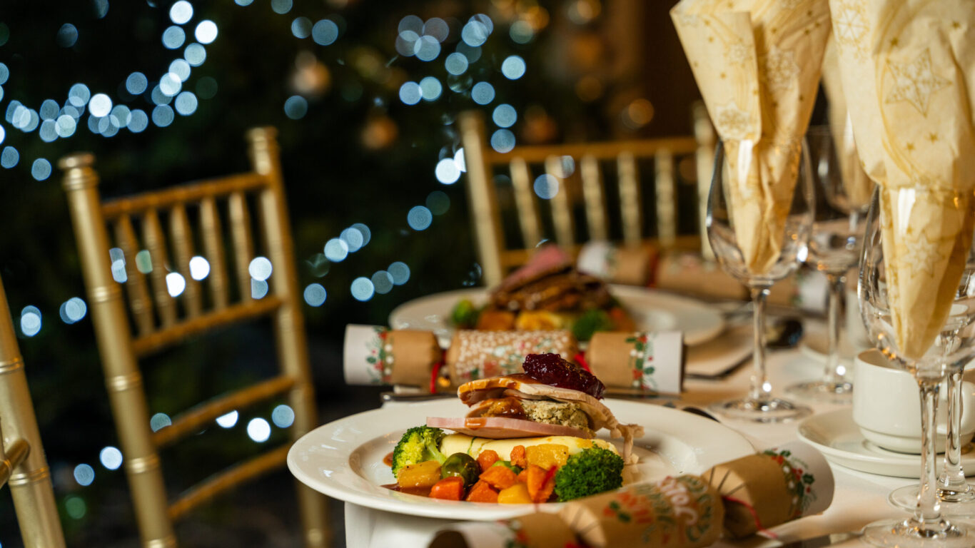 A close-up of a festive dining table set with plates of food, Christmas crackers, wine glasses, and a blurred Christmas tree with lights in the background.