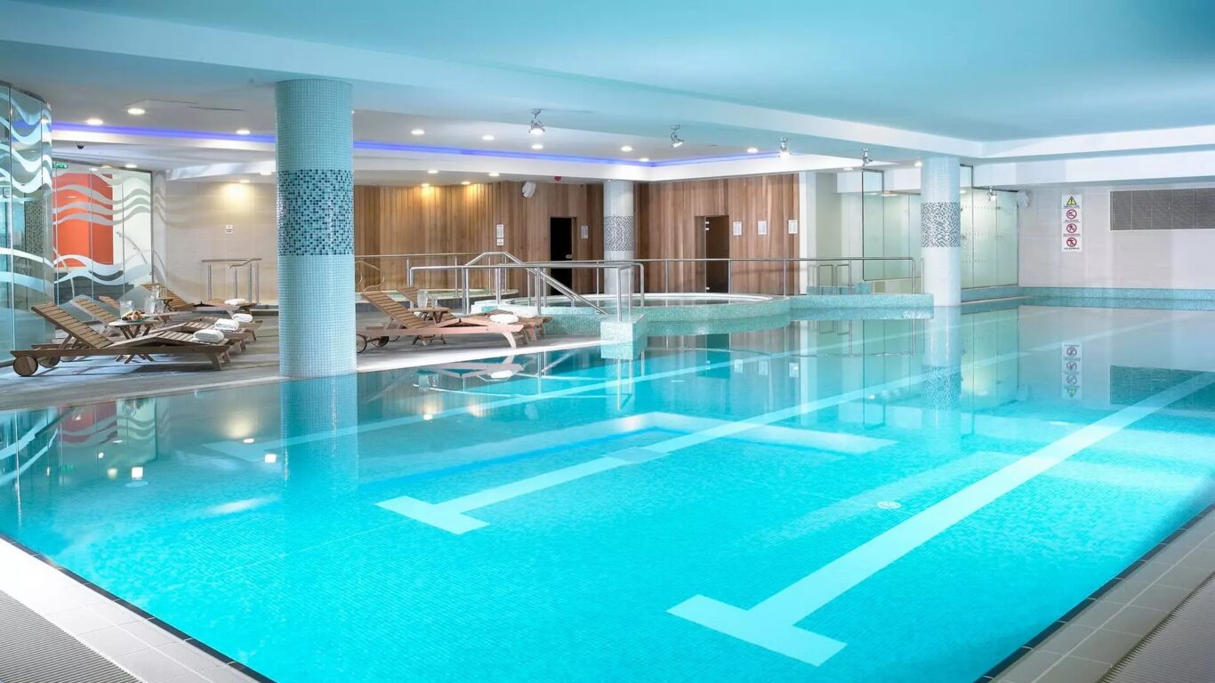 Enjoy a refreshing indoor swimming pool with clear blue water, poolside loungers, tiled pillars, and wood-paneled walls under bright ceiling lights—perfect for relaxing Spring Hotel Breaks.