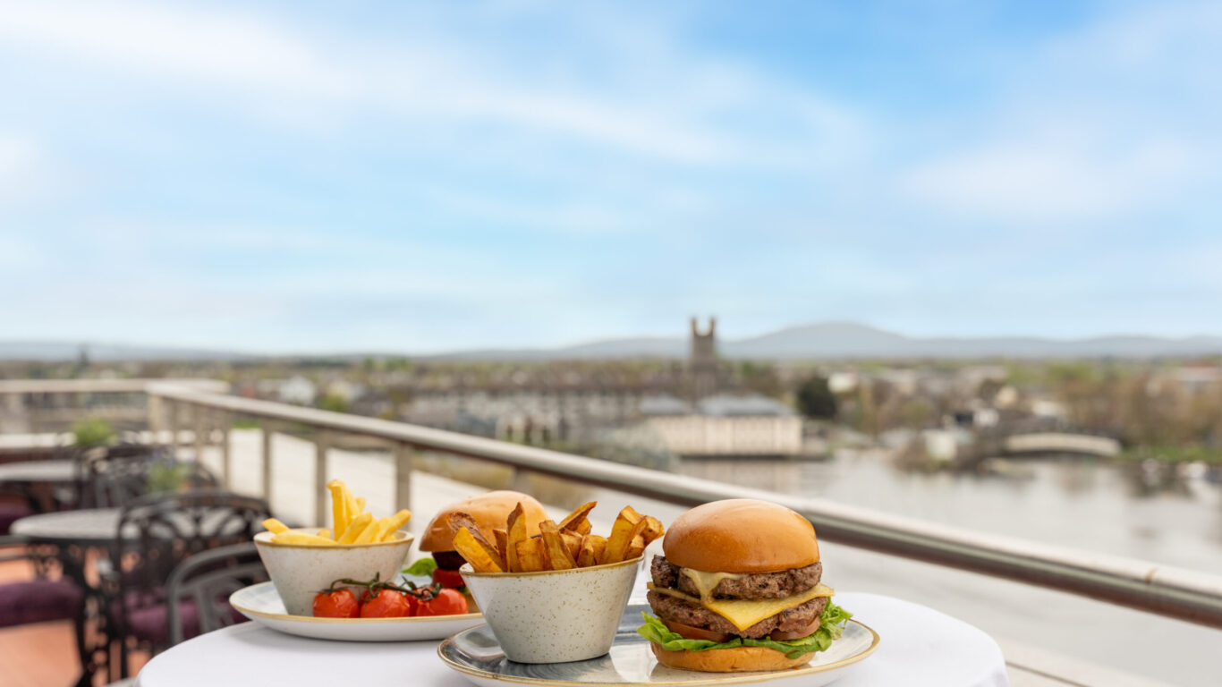 A plate with a cheeseburger, fries, and salad sits on a round table on an outdoor terrace overlooking a cityscape and river under a blue sky.