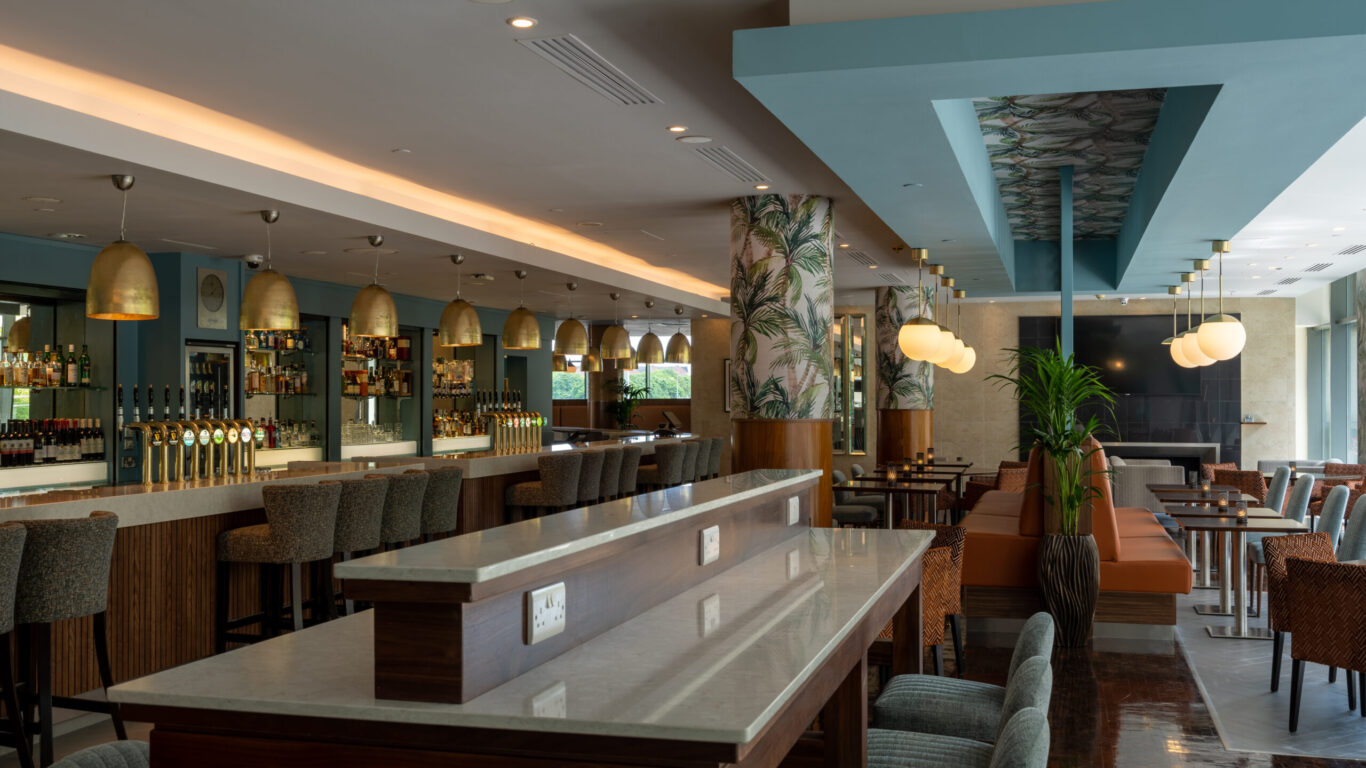 Modern bar and lounge area with high tables, upholstered chairs, a long bar counter, and pendant lighting, decorated with plants and large windows for natural light.