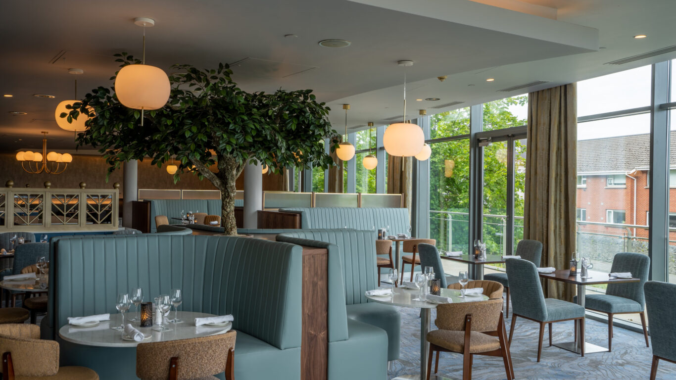 Modern restaurant interior with blue booths, wooden chairs, and round tables set for dining—an inviting space perfect for Spring Hotel Breaks, featuring large windows, an indoor tree, and elegant pendant lighting.