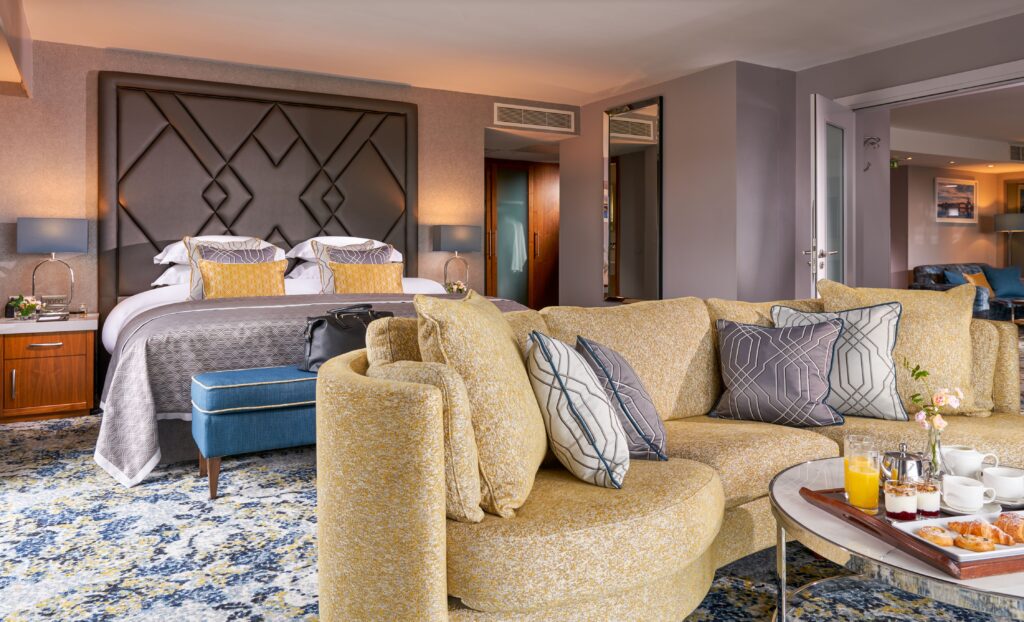 Modern hotel suite with a double bed, patterned pillows, a yellow curved sofa, coffee table with breakfast items, and contemporary decor throughout the room.
