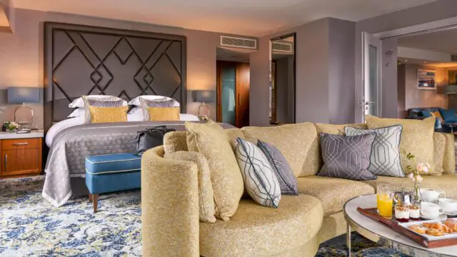Modern hotel suite with a double bed, patterned pillows, a yellow curved sofa, coffee table with breakfast items, and contemporary decor throughout the room.