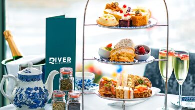 A table set for afternoon tea with a two-tier tray of pastries and sandwiches, a teapot, blue-patterned china, champagne glasses, and a River Restaurant menu by a window. Afternoon tea in Limerick