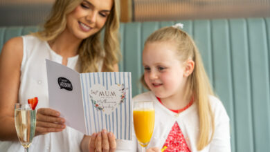 A woman and a young girl sit together in a booth, looking at a Mother&rsquo;s Day card. Two glasses of drinks are on the table in front of them.