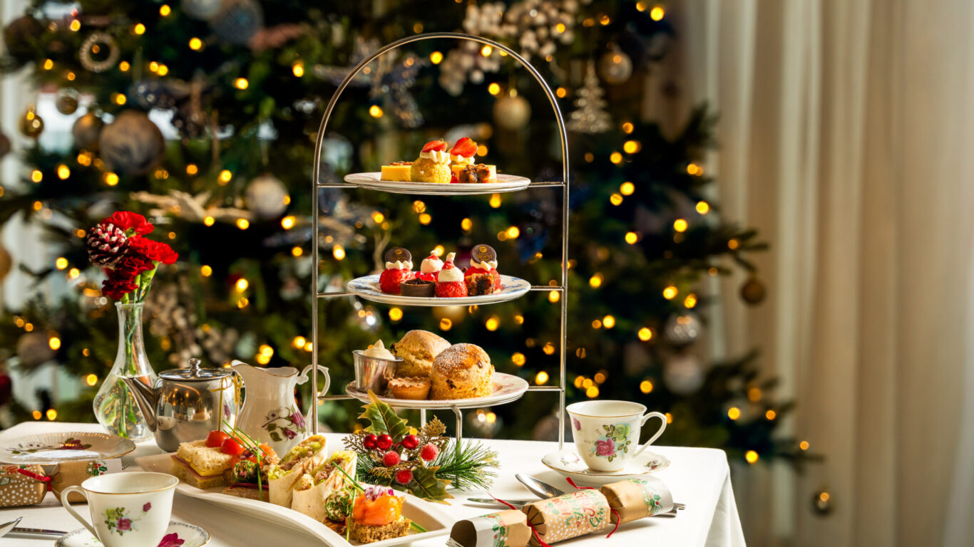 A table set for afternoon tea with assorted pastries and sandwiches, fine china, and a decorated Christmas tree with lights in the background.