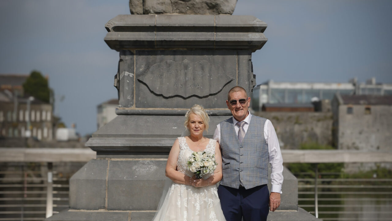 A bride in a white gown and a man in formal attire pose together in front of a stone monument on a sunny day.