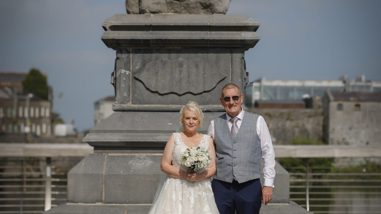 A bride in a white gown and a man in formal attire pose together in front of a stone monument on a sunny day.