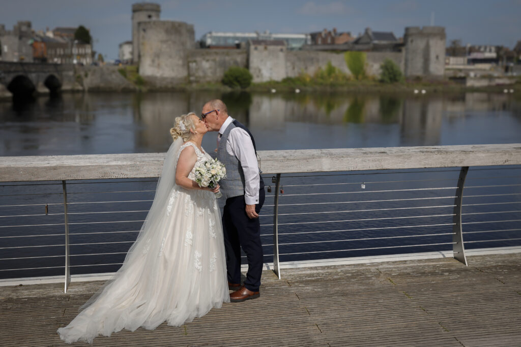 A bride and groom share a kiss on a riverside boardwalk with a historic stone castle and bridge in the background.