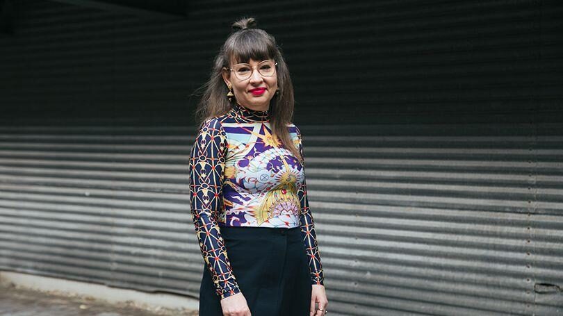 A woman with glasses and straight brown hair in a geometric-patterned long-sleeve top and black pants stands in front of a corrugated metal wall.
