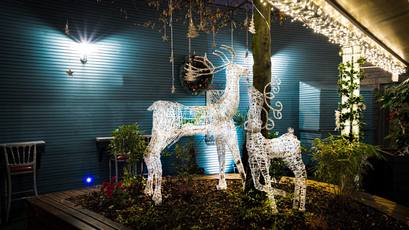 Two illuminated wireframe reindeer decorations stand under a tree in an outdoor area with string lights and a wreath on the wall.