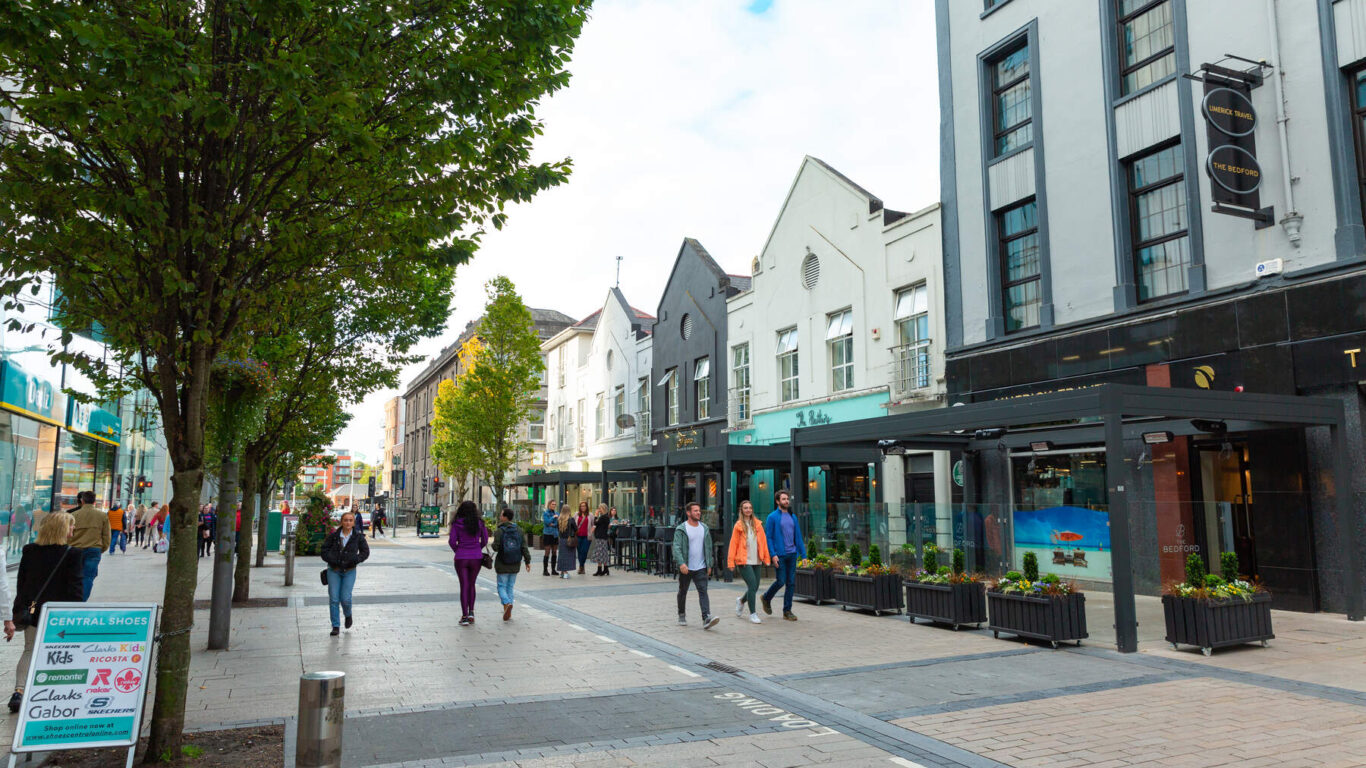 Pedestrians walk along a shopping street in Limerick City lined with trees, shops, and cafes, with outdoor seating visible and people interacting in a lively urban setting.