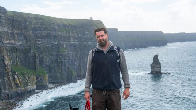 A man with a backpack stands on a grassy cliff edge holding a dog leash, with cliffs and the ocean visible in the background.