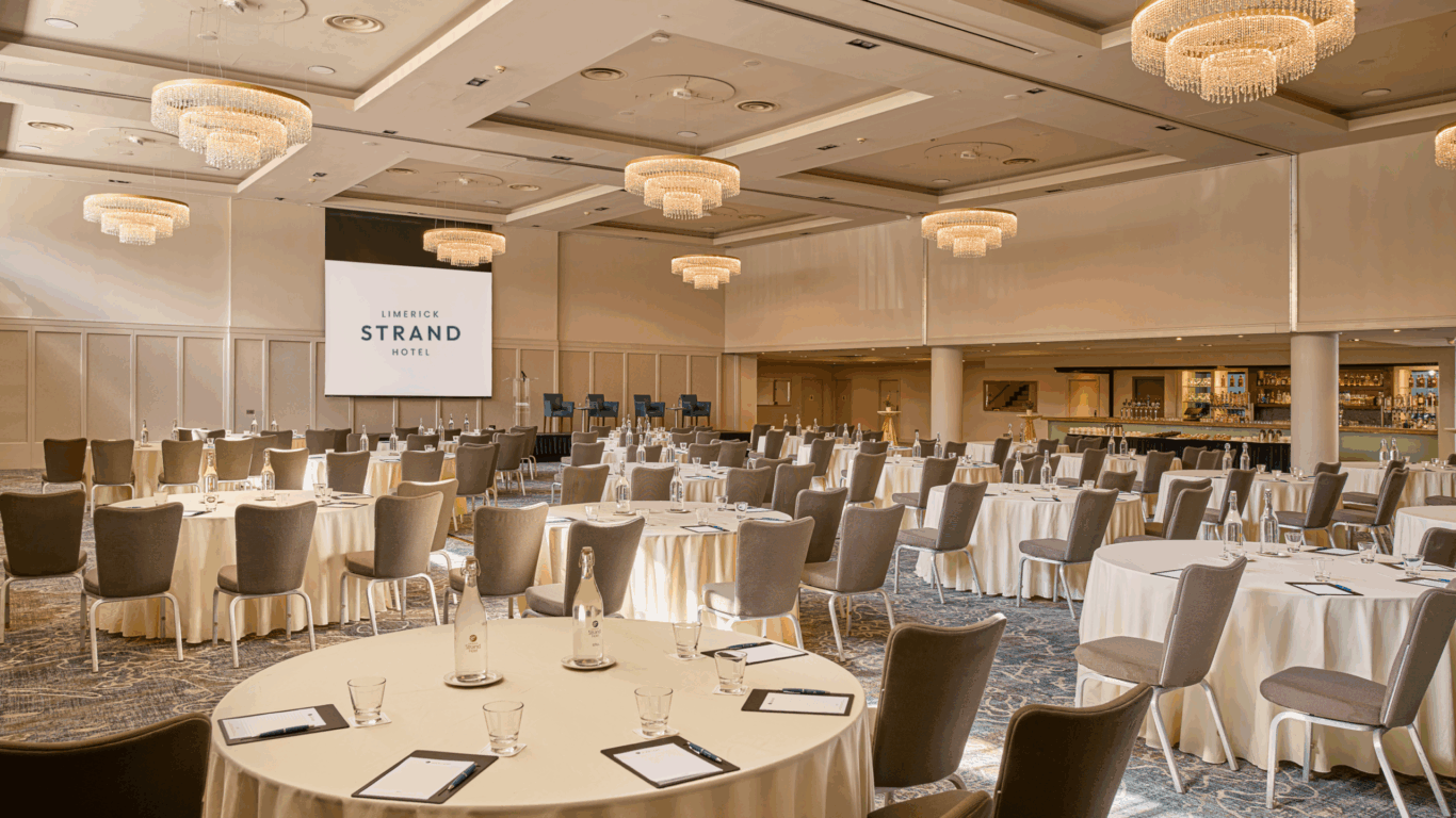 A spacious hotel conference room with round tables set for an event, a large projector screen, chandeliers, and a bar area in the background.
