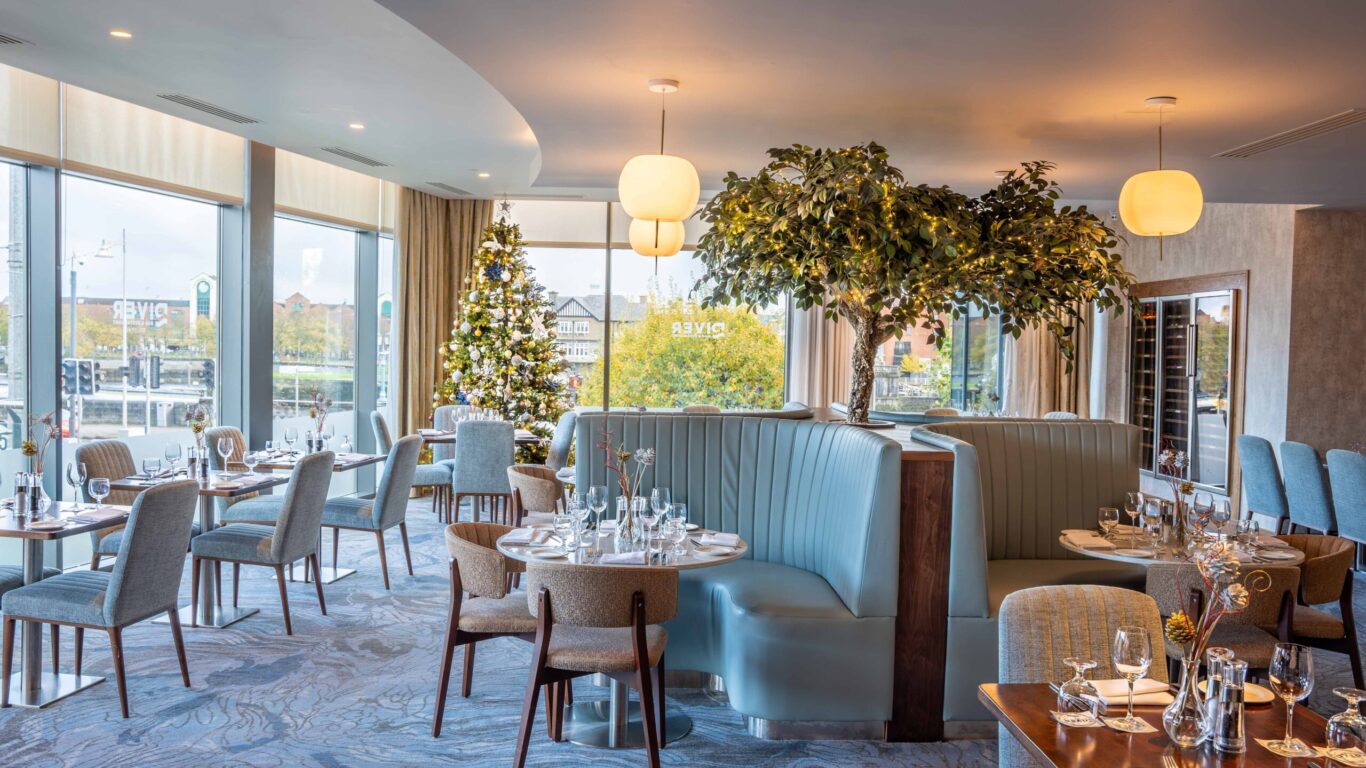 Modern restaurant interior with blue upholstered booths, set tables, large windows, indoor trees, and a decorated Christmas tree near the center.