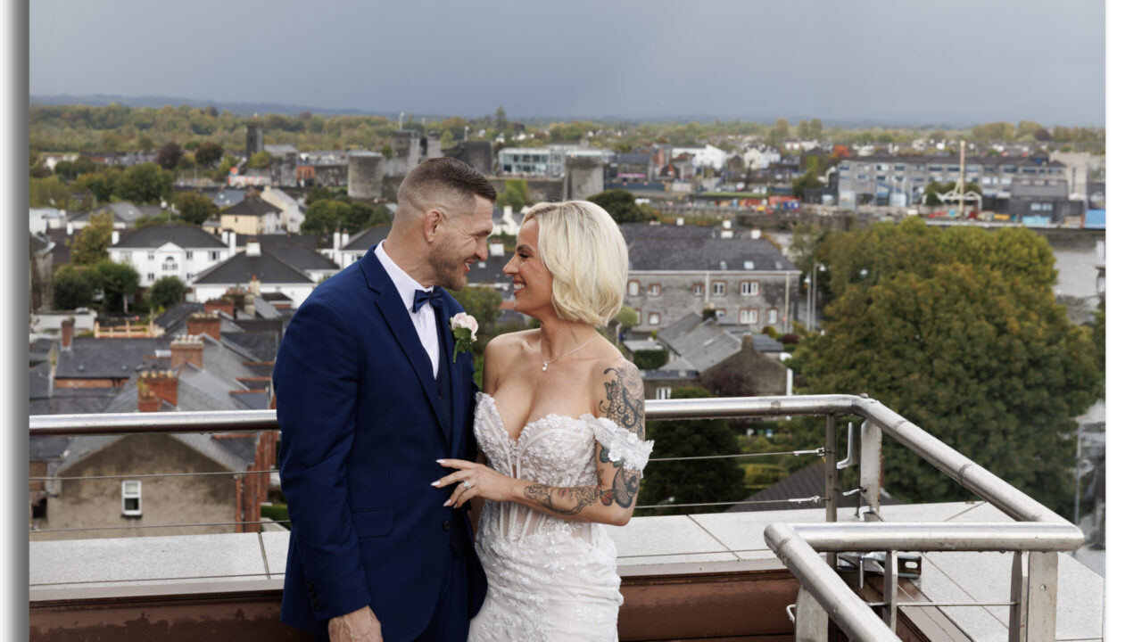 A bride and groom stand close together on a rooftop, smiling at each other, with a cityscape and overcast sky in the background.