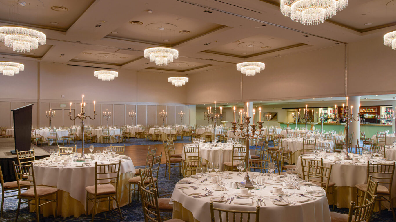 A formal banquet hall with round tables set for dining, gold chairs, candelabras, chandeliers overhead, and a bar area in the background.
