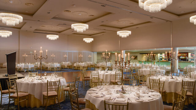A formal banquet hall with round tables set for dining, gold chairs, candelabras, chandeliers overhead, and a bar area in the background.