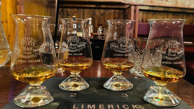 Four whiskey tasting glasses with amber liquid are arranged on a mat labeled