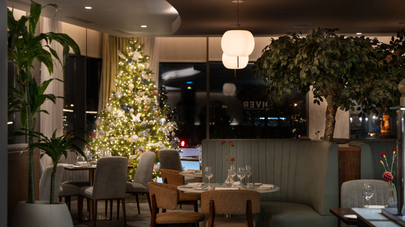 A modern restaurant dining area with neatly set tables, upholstered chairs, a decorated Christmas tree, and large indoor plants, softly lit in the evening.