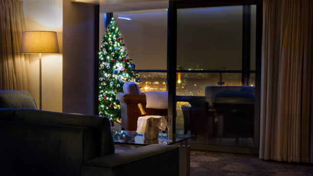 A living room at night with a lit Christmas tree by a large window, city lights outside, a sofa, a lamp, and a glass coffee table with a wine bottle and two glasses.