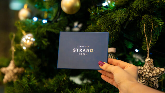 A hand holds a blue Limerick Strand Hotel envelope in front of a decorated Christmas tree with ornaments and lights.