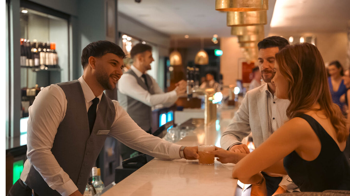 Bartender serving a drink to a seated woman at a modern bar, with other patrons and staff visible in the background.