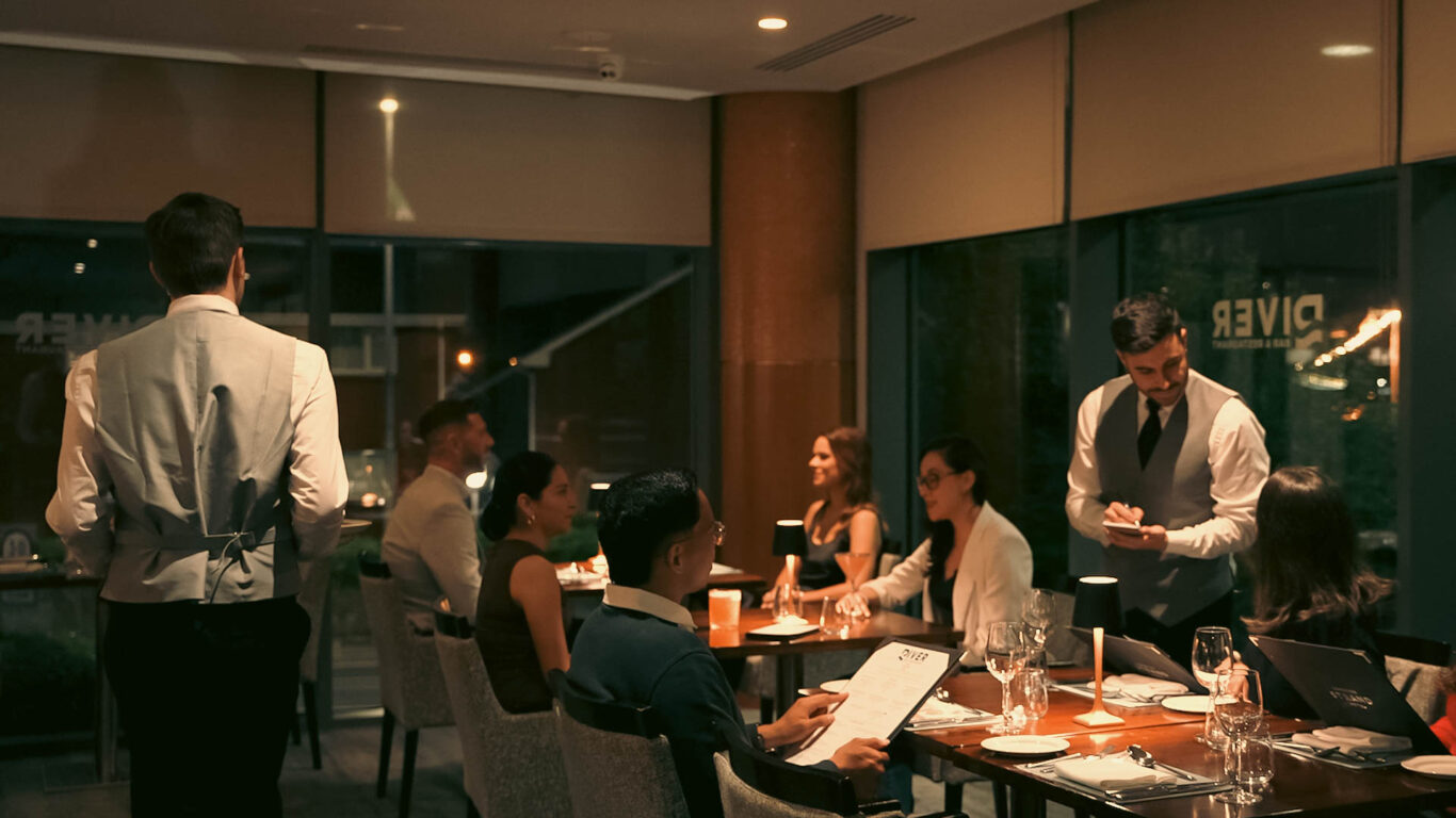Waiters serve dinner to seated guests in a dimly lit, modern restaurant with large windows and soft lighting.