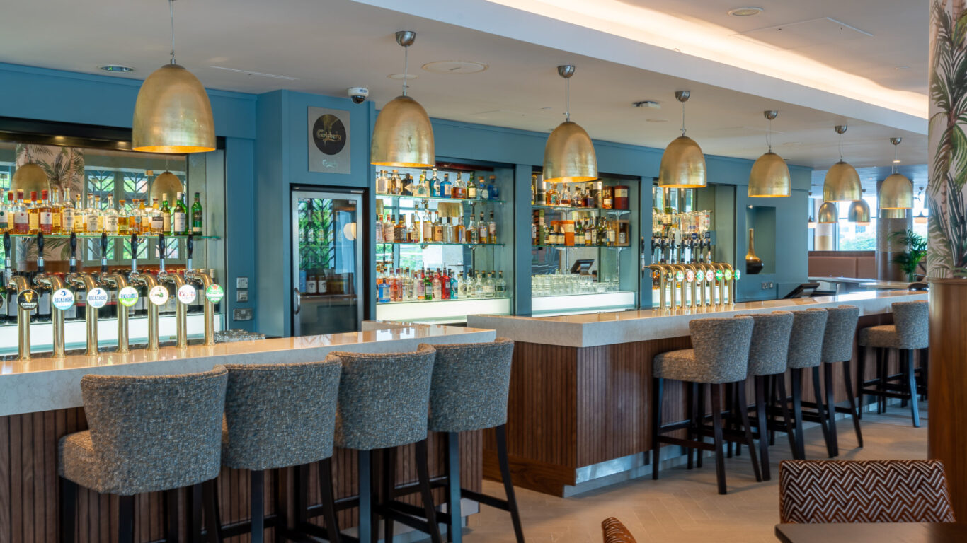 A modern bar with high upholstered stools, a marble countertop, rows of liquor bottles on shelves, and multiple hanging brass pendant lights.