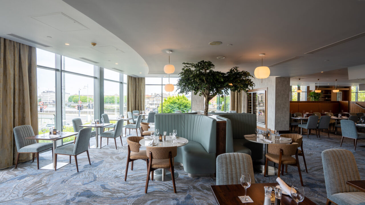 Modern restaurant interior with large windows, round tables, cushioned chairs, curved booth seating, and a tree feature in the center, set for service with glassware and napkins.