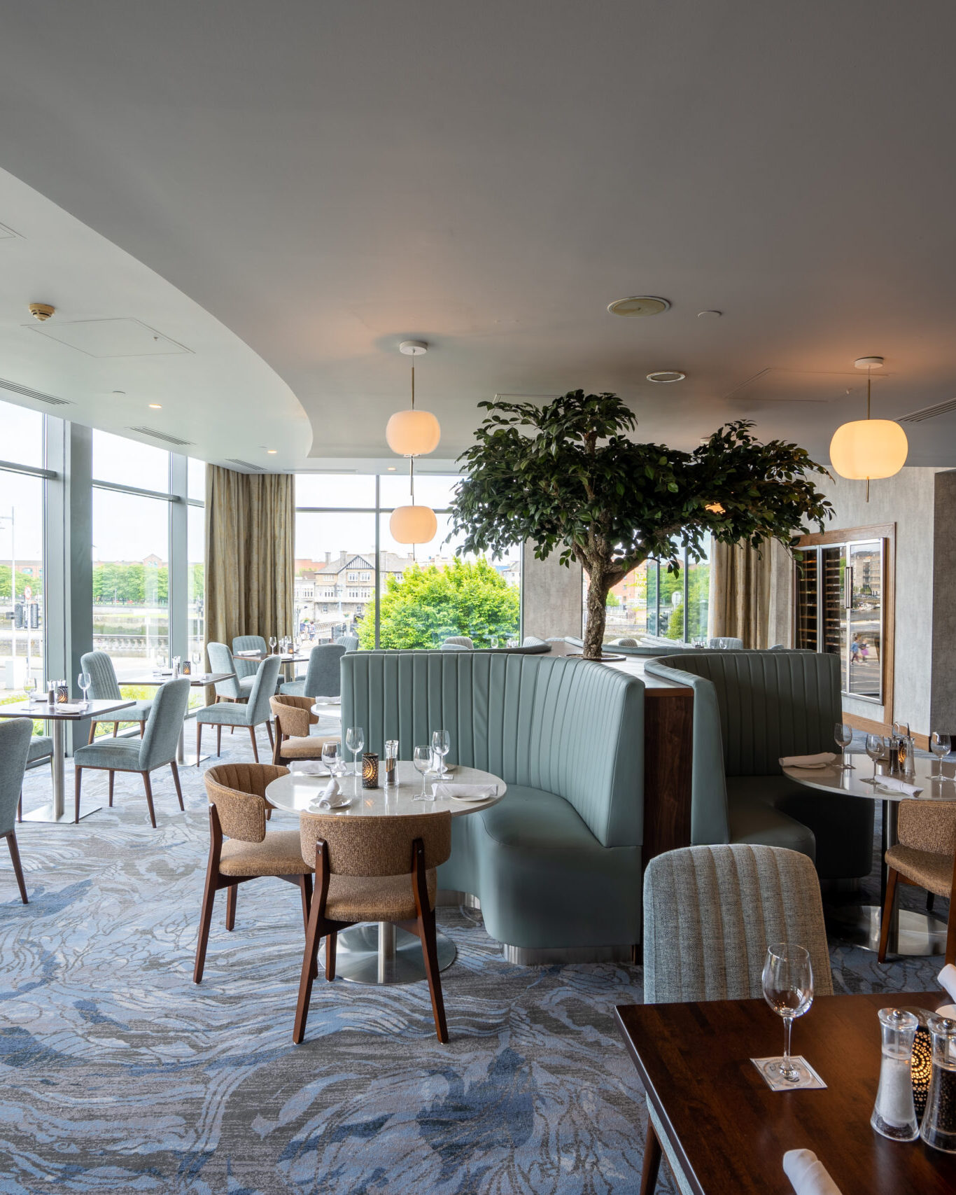 Modern restaurant interior with large windows, round tables, cushioned chairs, curved booth seating, and a tree feature in the center, set for service with glassware and napkins.