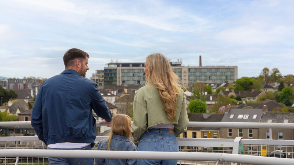 A man, woman, and child stand on a balcony overlooking a cityscape with buildings, trees, and a cloudy sky.