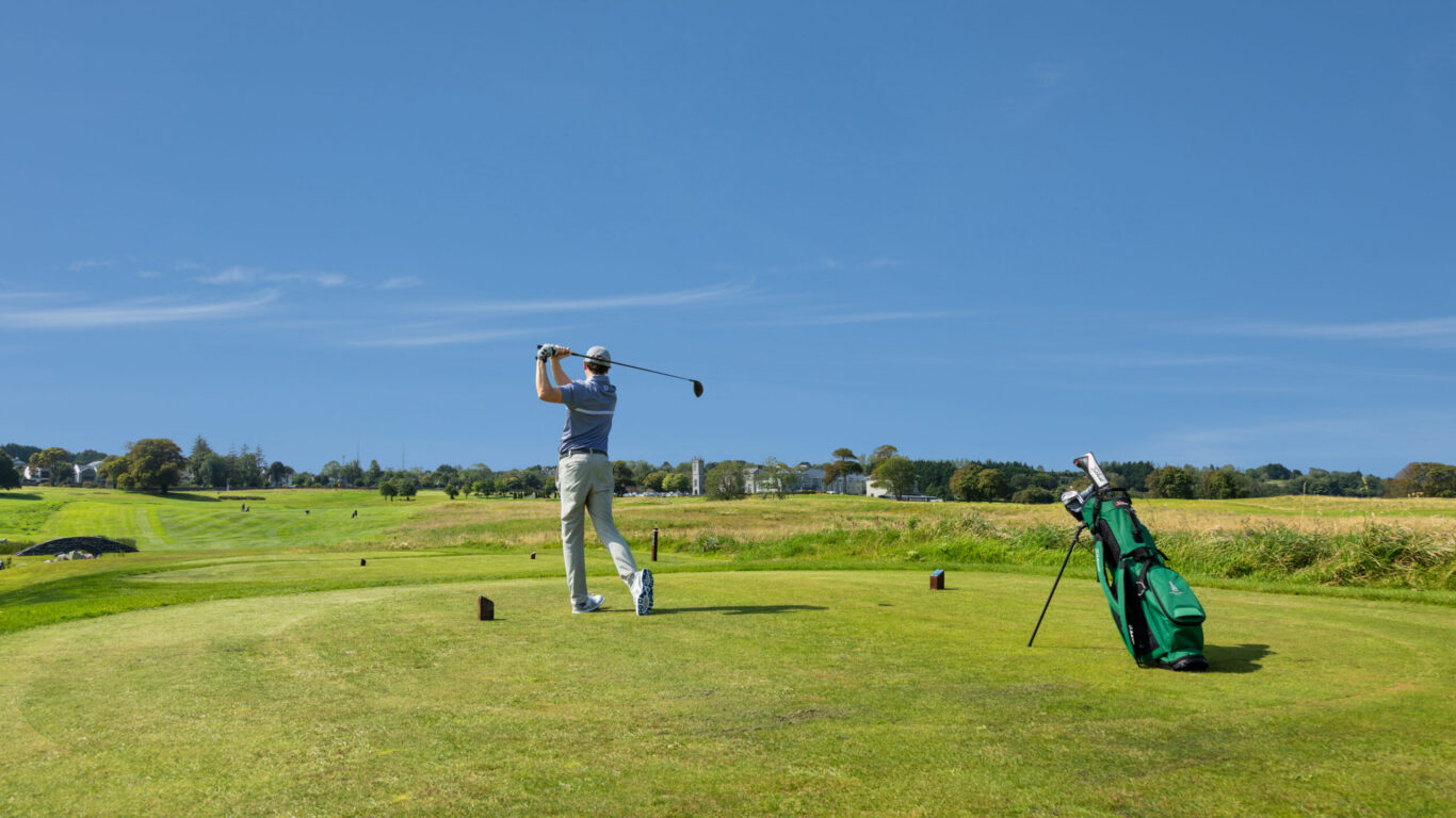 A golfer swings a club on a green golf course under a clear blue sky, with a golf bag standing nearby&mdash;perfect for those enjoying relaxing Autumn Hotel Breaks.