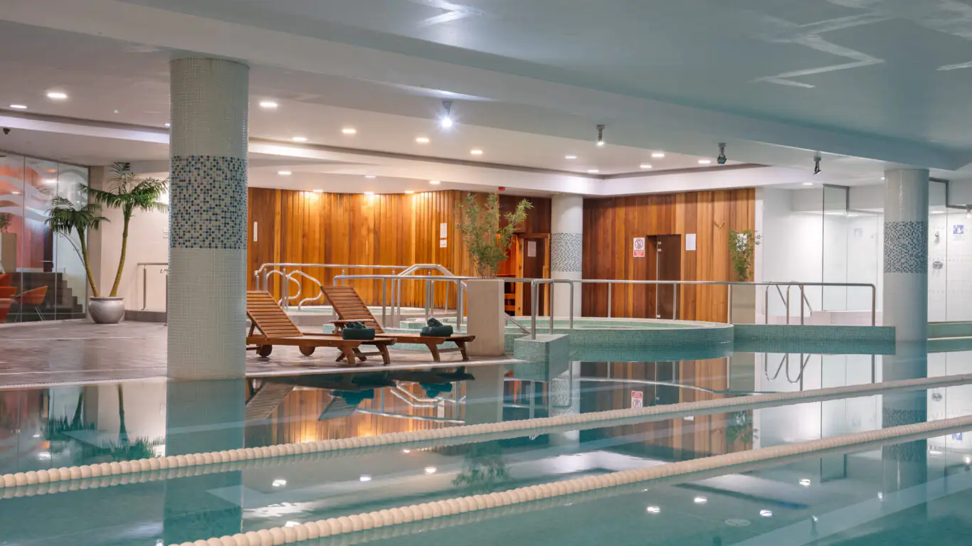 Indoor swimming pool area with wooden walls, two lounge chairs, potted plants, and handrails, featuring calm water and swimming lanes.