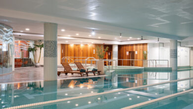 Indoor swimming pool area with wooden lounge chairs, a hot tub, tiled columns, and pool lane dividers, surrounded by wooden and glass walls and potted plants.