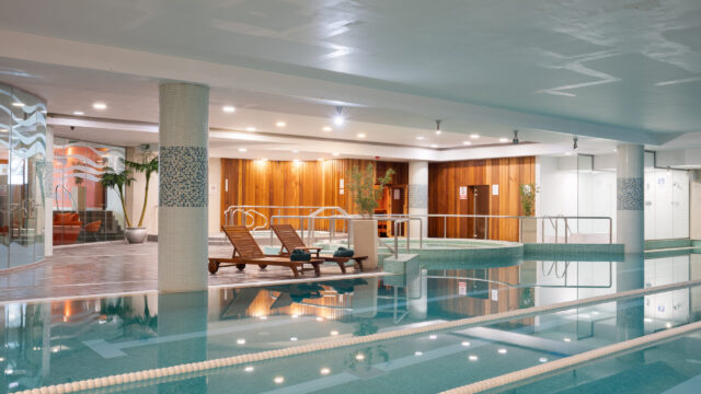 Indoor swimming pool area with wooden lounge chairs, a hot tub, tiled columns, and pool lane dividers, surrounded by wooden and glass walls and potted plants.