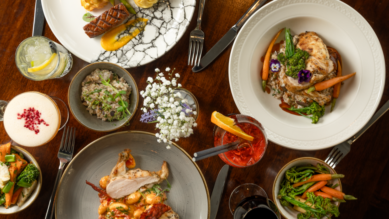 A table set with assorted gourmet dishes including grilled meat, vegetables, mashed potatoes, sauces, cocktails, and a small floral arrangement on a dark wooden surface.
