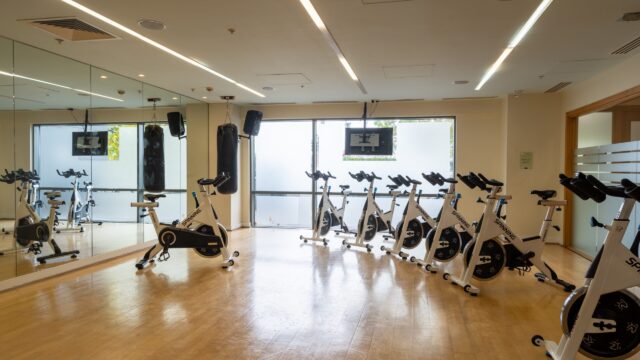 A bright indoor cycling studio with several stationary bikes lined up in front of mirrors and frosted windows; a TV is mounted on the back wall.