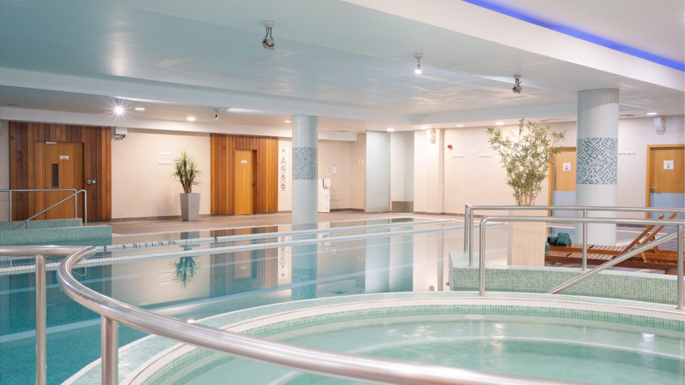 Indoor spa area with a circular hot tub in the foreground, a larger pool behind it, lounge chairs, plants, and wooden doors along the back wall.